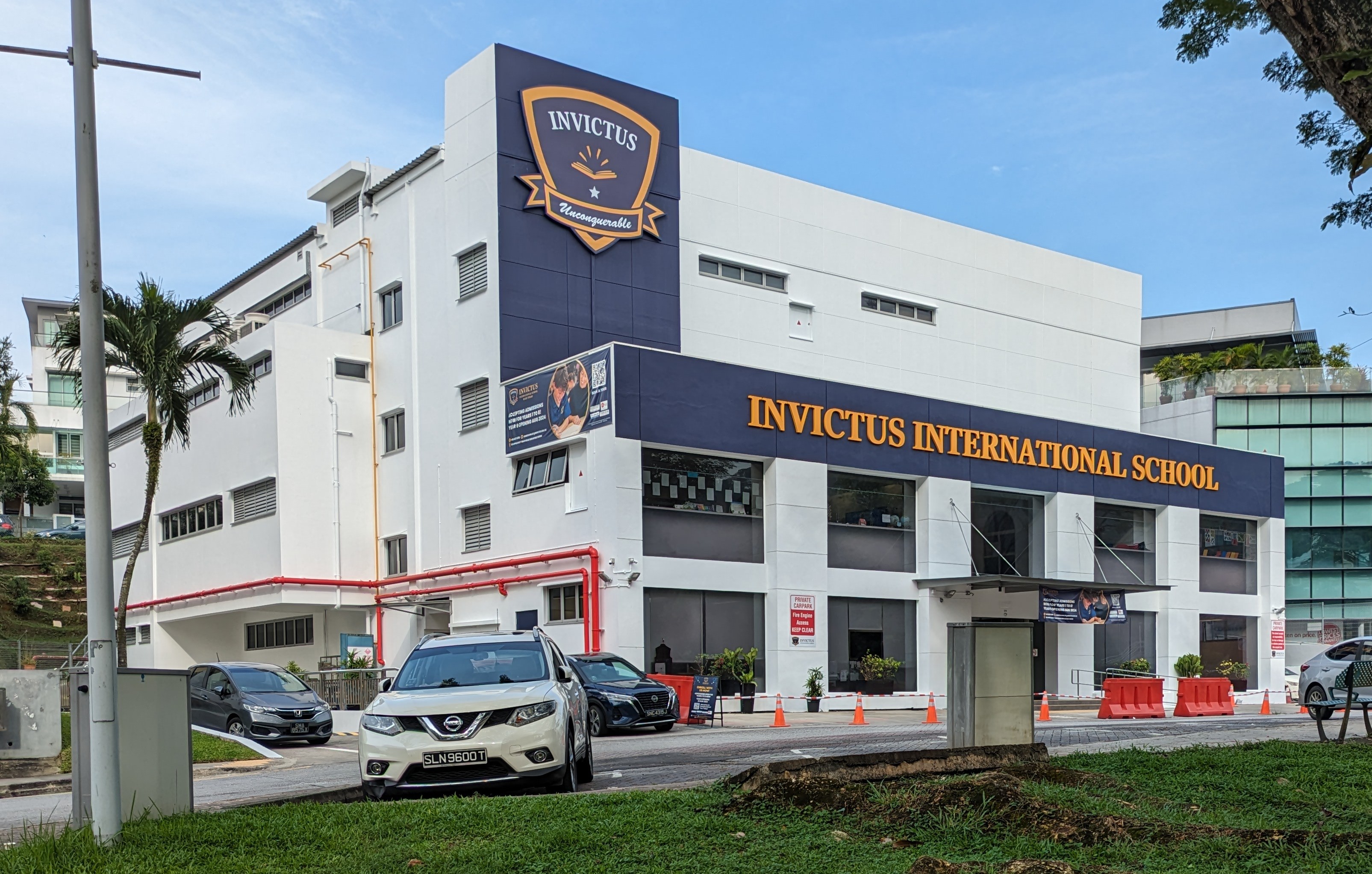 Exterior view of Invictus International School in Singapore, showing buildings, signage, vehicles, road infrastructure, greenery, and utility poles against a cloudy sky.
