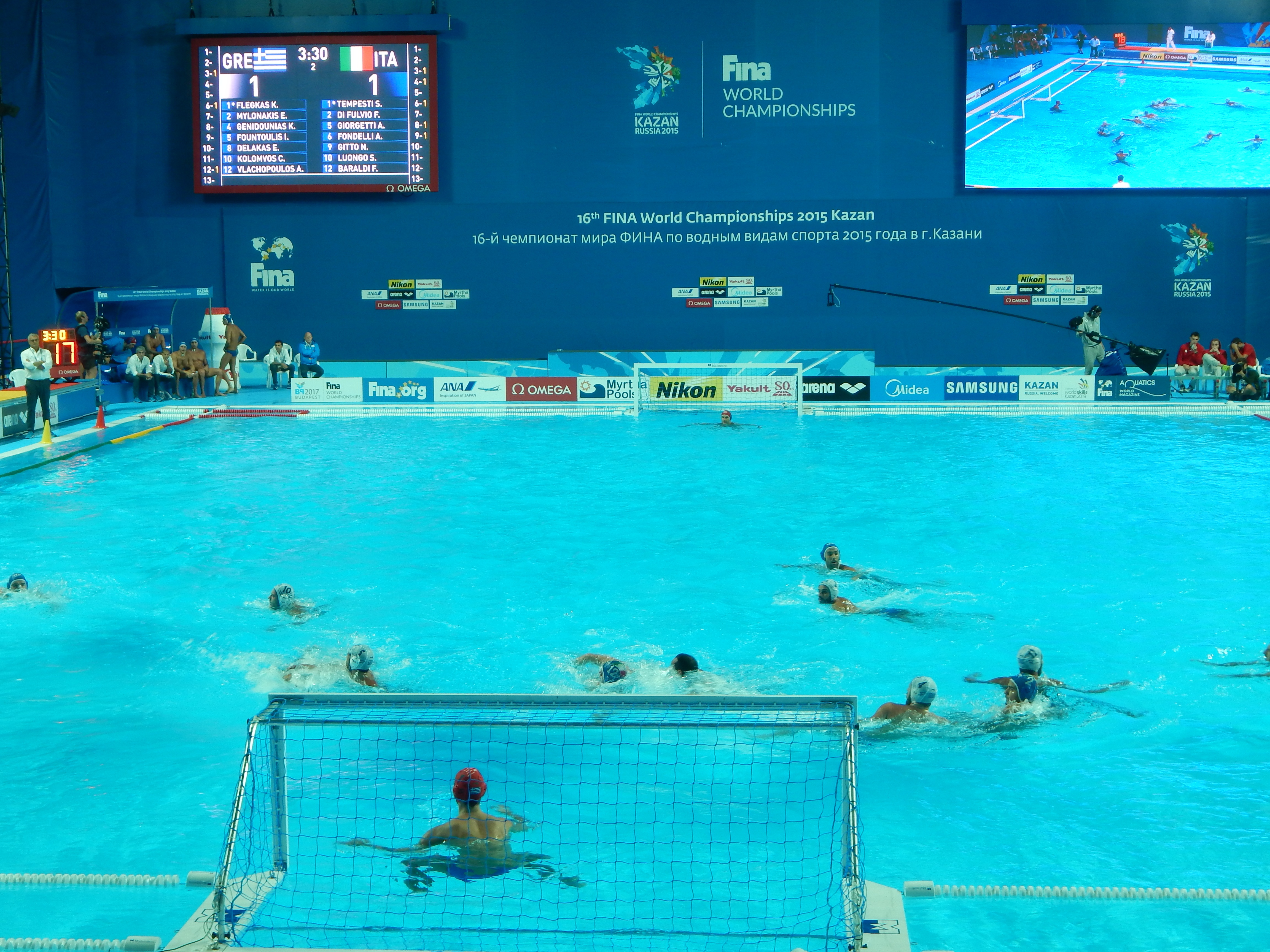 Eine Gruppe von Menschen spielt Wasserball in einem großen Pool mit einem Netz in der Mitte, Zuschauer links und ein Bildschirm mit Text und Logos im Hintergrund bei den 2016 FINA-Weltmeisterschaften in Kazan.