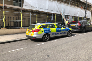 Ein Polizeiwagen steht auf der Straße neben einem Gebäude, daneben stehen zwei Helme tragende Personen; im Vordergrund sind ein Geländer und ein Pfahl zu sehen, und im Hintergrund hängt ein weißes Tuch über das Gebäude, was auf eine Polizeiermittlung hindeutet.