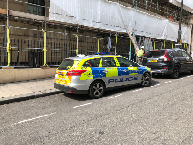Ein Polizeiwagen steht auf der Straße neben einem Gebäude, daneben stehen zwei Helme tragende Personen; im Vordergrund sind ein Geländer und ein Pfahl zu sehen, und im Hintergrund hängt ein weißes Tuch über das Gebäude, was auf eine Polizeiermittlung hindeutet.