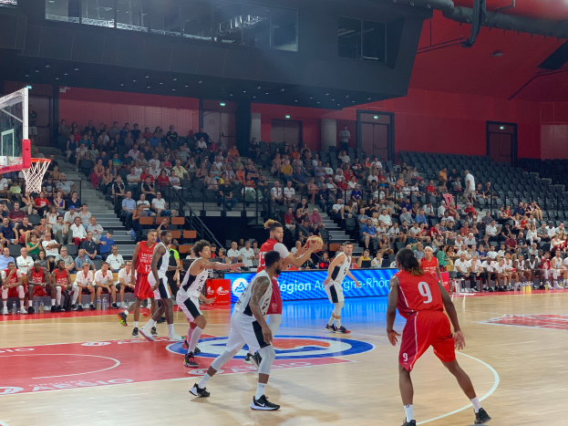 Eine Gruppe von Männern spielt Basketball in einer Turnhalle mit einem Netz auf der linken Seite, Zuschauer sitzen im Hintergrund und Scheinwerfer erhellen die Szene von oben.