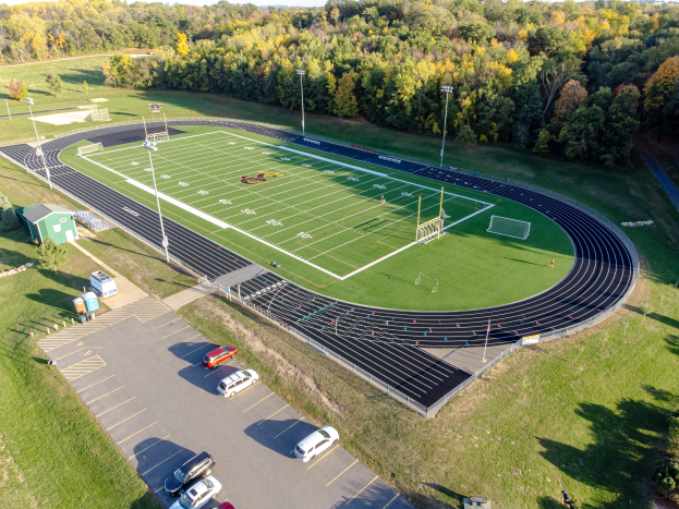 Eine Luftaufnahme eines Fuballfelds mit geparkten Autos auf dem Parkplatz, umgeben von grünem Gras, Bäumen, Laternenmästen und Häusern, unter einem klaren Himmel.