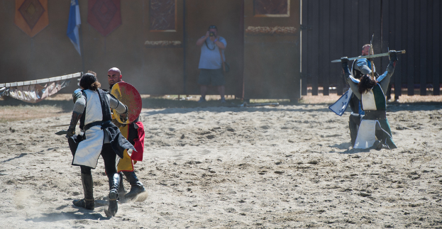 Eine Person steht in der Mitte und hält eine Kamera, um eine Szene von Menschen zu filmen, die auf beiden Seiten kämpfen, mit einer Wand, einem Tor und einer Flagge im Hintergrund und Sand unten.