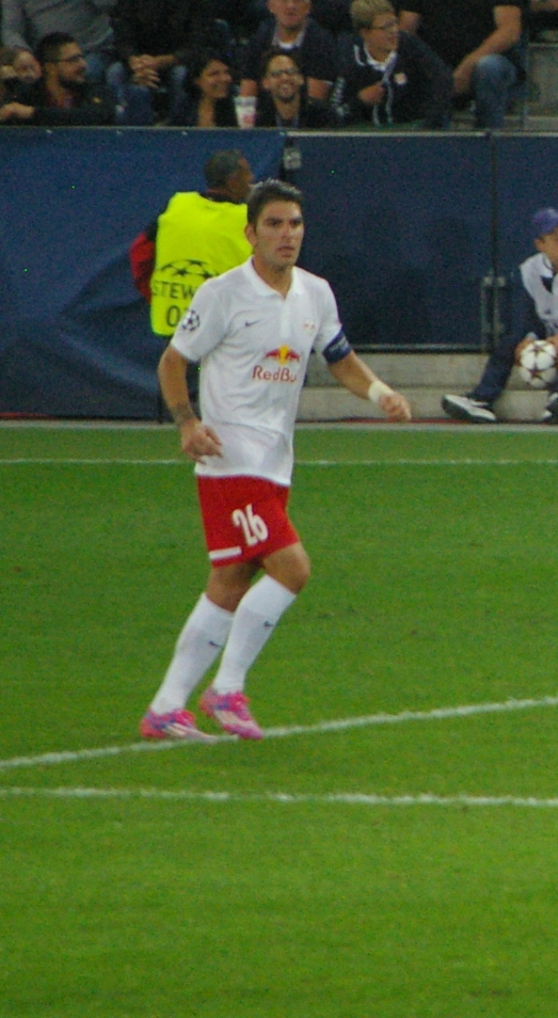 Ein Mann in einem weißen Hemd und roten Shorts spielt Fußball auf einem Feld, umgeben von einer Gruppe von Menschen im Hintergrund.