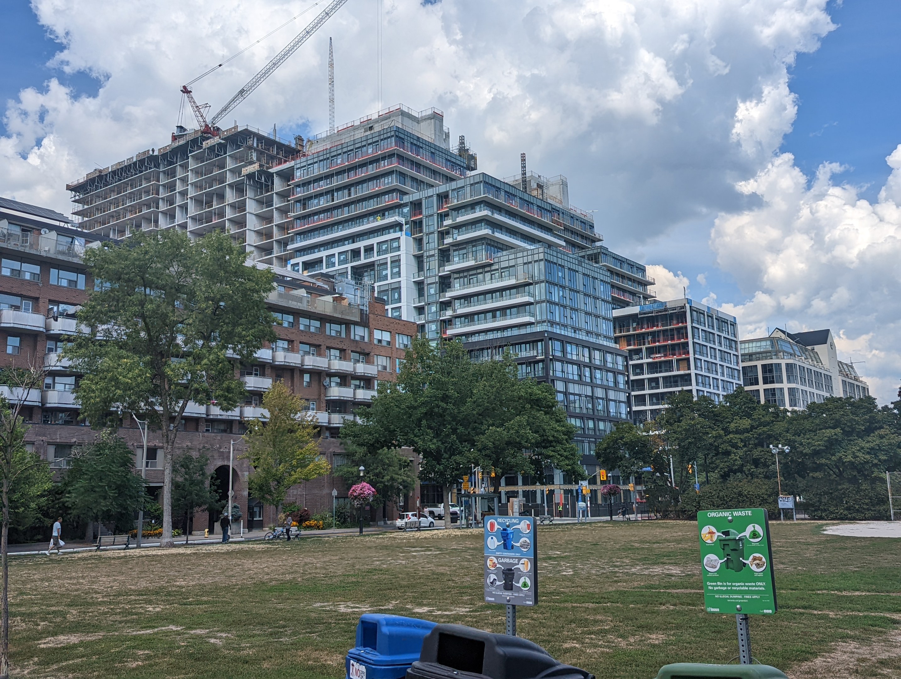 Large building under construction in a park, identified as the Toronto Housing Authority, surrounded by construction materials, trees, and people, with a blue and white sky.