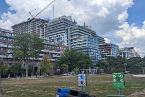 Large building under construction in a park, identified as the Toronto Housing Authority, surrounded by construction materials, trees, and people, with a blue and white sky.