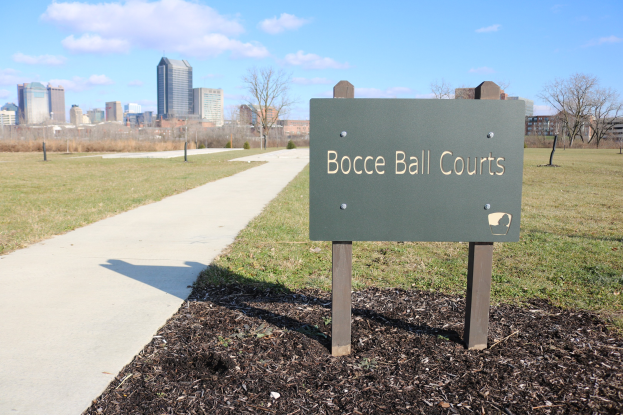 Ein Schild am Boden in der Nähe eines Weges und Bäume mit der Aufschrift "Bocce Ball Courts in Indianapolis, Indiana" und einem bewölkten Himmel im Hintergrund.