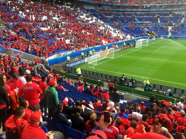 Eine große Menschenmenge in einem Stadion schaut ein Fußballspiel zu, mit Sitzplätzen und Stehplätzen, Bannern, einem Torpfosten und Deckenleuchten.