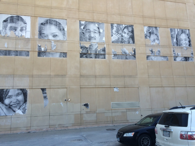 Ein GebÃ¤ude mit Autos und Gullis auf der StraÃŸe im Vordergrund und Plakate von Menschen an einer Wand im Hintergrund.