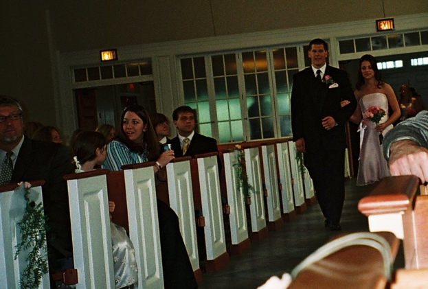 Eine Gruppe von Menschen sitzt auf einer Bank, während eine Braut und ein Bräutigam einen Weg entlang auf sie zukommen, die Braut hält einen Strauß Blumen, mit Laternen und einem Fenster im Hintergrund, das eine Kirchenatmosphäre suggeriert.