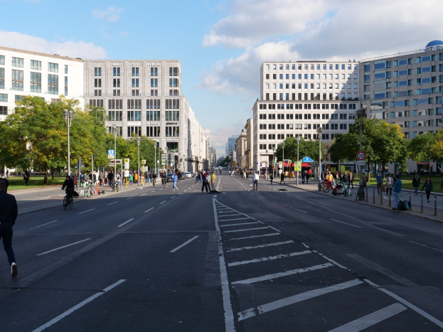 Eine belebte Straßenkreuzung in Berlin, Deutschland, mit Fußgängern und Radfahrern auf der Straße, hohen Gebäuden, Bäumen, Laternenmasten und Schildern, unter einem bewölkten Himmel.