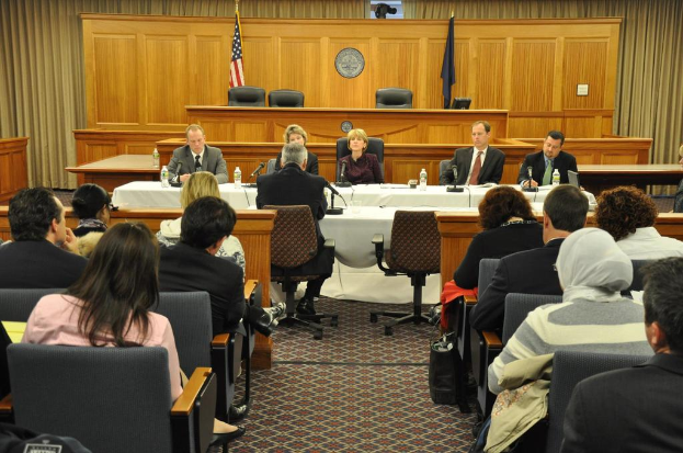 Ein Versammlungsraum mit einer Gruppe von Menschen, die auf Stühlen sitzen, zwei ausländischen Flaggen, einem orangefarbenen Tisch mit Wasserflaschen und einem Mikrofon.