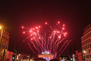 Eine Stadtstraße in Berlin an Silvester, voller Gebäude, Bäume, Laternenpfähle, Ampeln, Schilder, Zelte und Menschen, mit einem von Feuerwerk erleuchteten Himmel im Hintergrund.