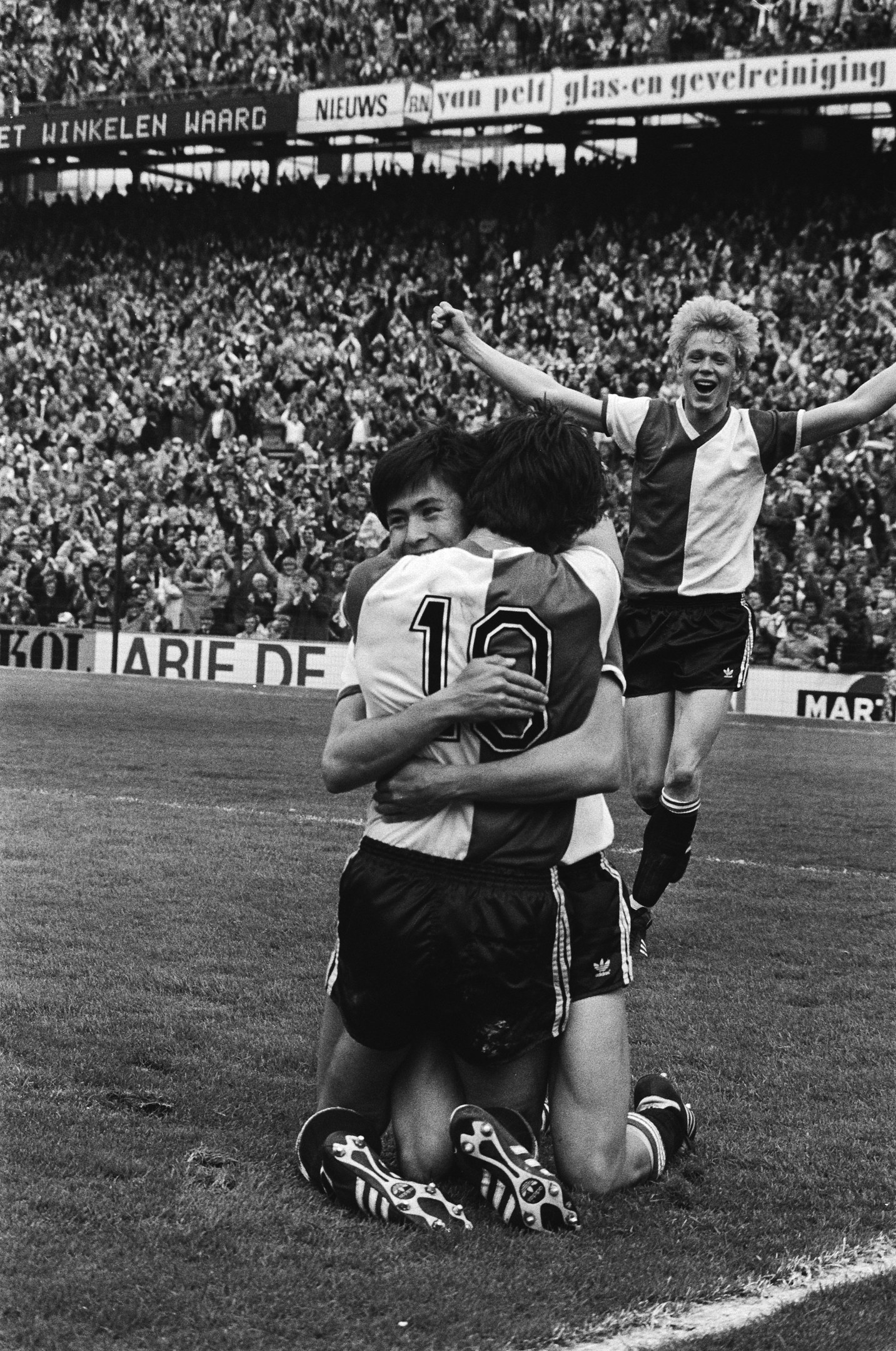 Drei Fußballspieler feiern ein Tor, indem sie sich auf dem Boden umarmen, vor einem jubelnden Publikum und schwenkenden Fahnen in einem schwarz-weißen Stadion.
