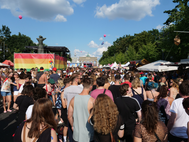Eine große Menschenmenge, die eine Straße mit Zelten, Bäumen, Pfählen, Laternen und Gebäuden im Hintergrund entlanggeht, bei der Berlin Pride Festival.
