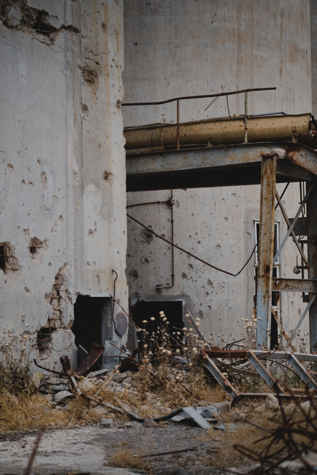 Altes Gebäude mit bröckelnden Wänden, kaputten Fenstern und einer zentralen Metallstruktur umgeben von Pflanzen, Eisenstangen und verstreuten Trümmern.