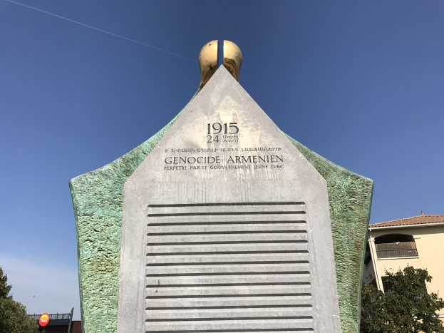 Ein steinernes Denkmal mit einer Uhr darauf, das mit Text beschriftet ist, steht in einem Park umgeben von Bäumen und Gebäuden unter einem klaren Himmel, zum Gedenken an den 24. Jahrestag des Völkermords in Armenien.
