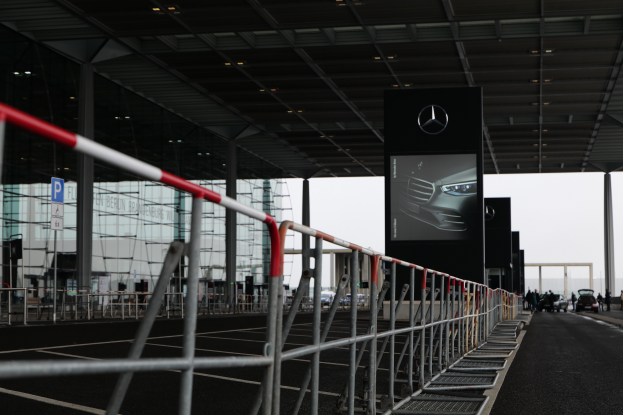 Eine Mercedes-Benz-Ausstellung auf der IAA in Frankfurt mit Fahrzeugen, Geländern, Pfählen, Schildern, Lichtern und einem Gebäude im Hintergrund, bei dem der Himmel oben zu sehen ist.