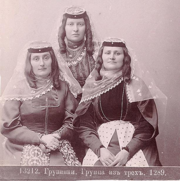 Drei Frauen in traditionellen russischen Hochzeitskleidern mit aufwändiger Stickerei und Perlen stehen vor einer weißen Wand, mit Text unten, der darauf hinweist, dass es ein russisches Hochzeitsfoto ist.