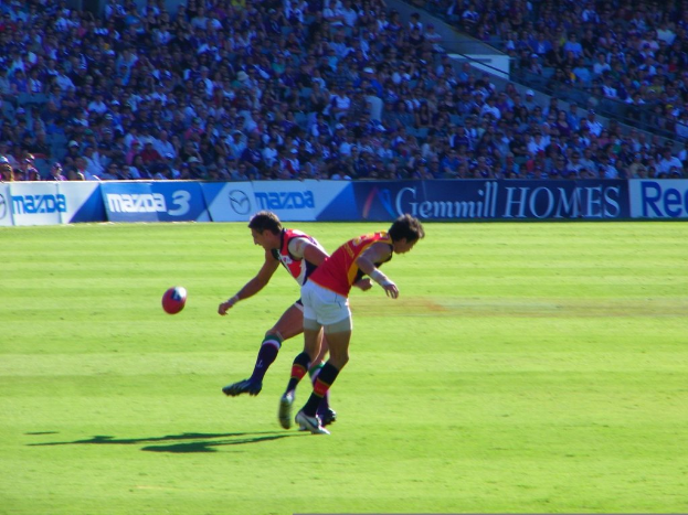 Zwei Personen in rot-weißer Kleidung stehen mit einem Ball in der Luft, während im Hintergrund des Stadions eine Menge und Banner zu sehen sind.