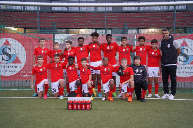 Eine Gruppe junger Männer in roten T-Shirts und schwarzen Shorts steht auf einem Fußballfeld vor einer Kiste roter Gegenstände, im Hintergrund sind Schilder mit der Aufschrift "FC Rot-Schwarz Eglaute - Champions League" zu sehen.