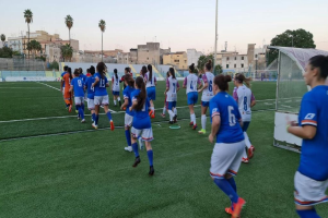 Eine Gruppe von Frauen, die auf einem Feld mit Gebäuden, Bäumen und Pfählen unter einem klaren blauen Himmel Fußball spielen, mit einem Torpfosten und Netz auf der rechten Seite.