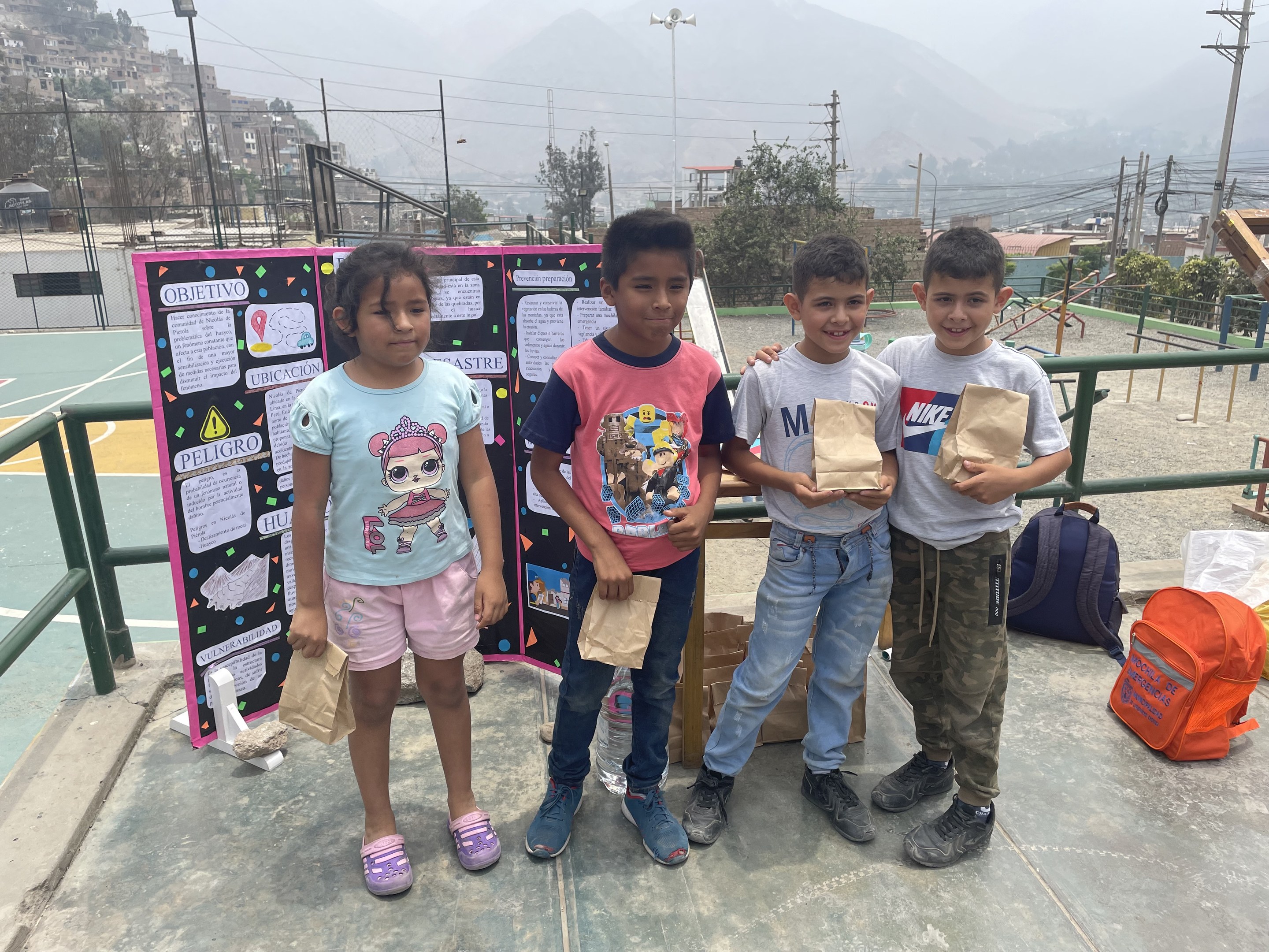 Gruppe von Kindern auf einem Basketballfeld mit Papiertüten in den Händen, mit Tüten und Gegenständen auf der rechten Seite und einer Tafel mit Text auf der linken Seite, vor einem Hintergrund aus Zäunung, Straßenlaternen, Bäumen, Gebäuden, Höhen und Himmel.