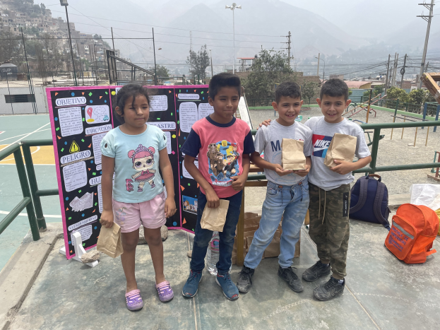 Gruppe von Kindern auf einem Basketballfeld mit Papiertüten in den Händen, mit Tüten und Gegenständen auf der rechten Seite und einer Tafel mit Text auf der linken Seite, vor einem Hintergrund aus Zäunung, Straßenlaternen, Bäumen, Gebäuden, Höhen und Himmel.