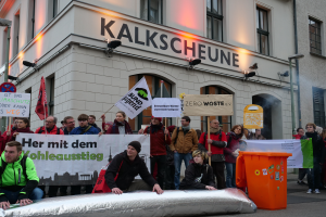 Eine Gruppe von Menschen mit Schildern und Plakaten steht vor einem Geb√§ude, mit zwei Personen im Vordergrund und einem M√ľllcontainer auf der rechten Seite, w√§hrend einer Demonstration in Deutschland, mit Geb√§uden, Fenstern, Lichtern und Schildern im Hintergrund.