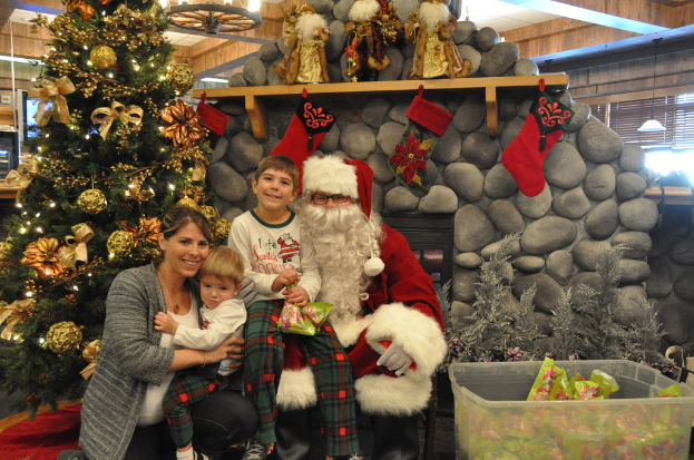 Eine Frau, Kinder und ein Mann in Santa-Klaus-Kostüm mit Brille und Handschuhen stehen neben einem geschmückten Weihnachtsbaum, einer Wand mit Felsen, Spielzeugen, Dekorationen, einem Korb mit Gegenständen und Hintergrundbeleuchtung.