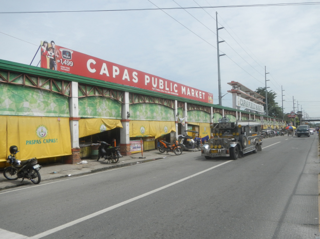 A bustling city street with vehicles, a footpath, electric poles, buildings, trees, and a cloudy sky, featuring a building with a "Capas Public Market" sign in the foreground.