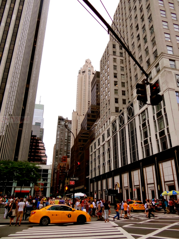 Eine Stadtstraße mit Gebäuden, Ampeln, Fenstern, einigen Passanten, Autos und Himmel.