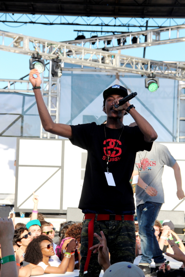 Eine Person in einem schwarzen T-Shirt h├Ąlt ein Mikrofon auf einer B├╝hne mit Menschen drumherum, w├Ąhrend eine andere Person in einem grauen T-Shirt im Hintergrund unter einem blauen Himmel steht.