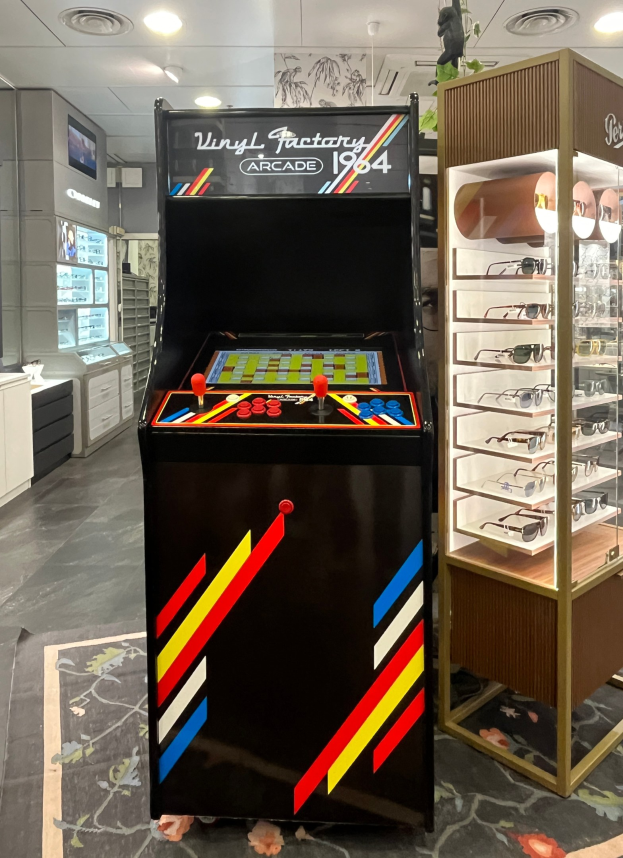 Ein Geschäft mit einem zentralen Retro-Arcade-Automaten, einer Glasvitrine auf der rechten Seite und einer retro-stylischen Beleuchtung mit Schränken, einem Fernseher und anderen Gegenständen im Hintergrund.
