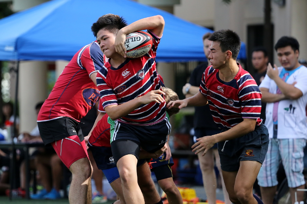 Eine Gruppe von Menschen, die draußen ein Spiel spielen, mit einem Jungen in der Mitte, der einen Ball hält, während andere daneben stehen und zuschauen, im Hintergrund ein blaues Zelt.