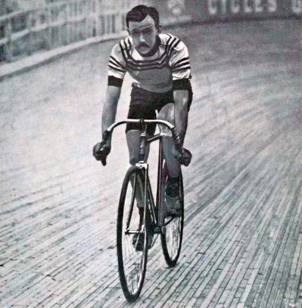 Schwarz-weißes Foto eines Mannes, der auf einer Bahn radfährt, mit einem Zaun und einer Tafel im Hintergrund.