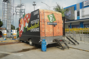 Ein großer Lkw steht vor einem Gebäude mit einer Tafel, auf der "Teilen Sie eine positiv überladene Erfahrung" steht, mit Stufen, einem Zylinder auf dem Boden, Geländern, Strommasten, Laternen, Bäumen und einem bewölkten Himmel.