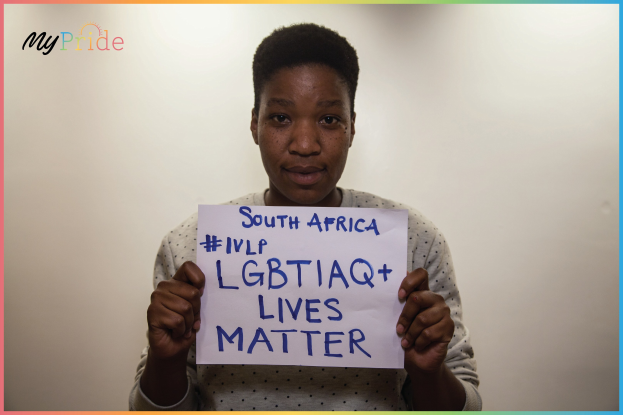 A woman in a white shirt stands determinedly in front of a wall, holding a sign that reads "LGBTQ+ Lives Matter" in bold black letters.