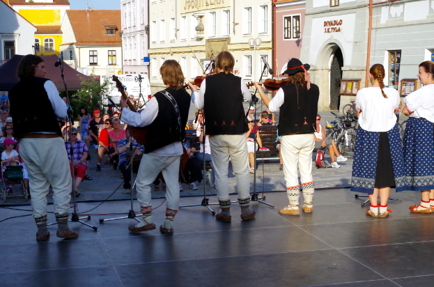 Vier Personen, die Geige spielen, auf einer Bühne mit einigen Zuschauern davor, Gebäuden im Hintergrund und zwei Frauen in der rechten Ecke.