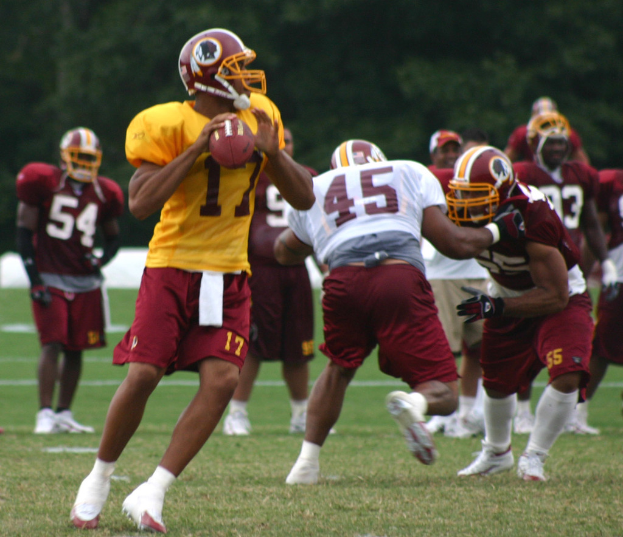 Eine Gruppe von Menschen in Jerseys, Helmen und weißen Schuhen spielt amerikanisches Football auf einem grasbewachsenen Feld mit Bäumen im Hintergrund.