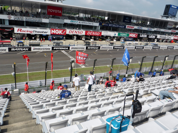 Eine Gruppe von Menschen sitzt auf den Tribünen einer Rennstrecke, umgeben von Fahnen, Texttafeln, Geländern und Stufen, unter einem bewölkten Himmel.