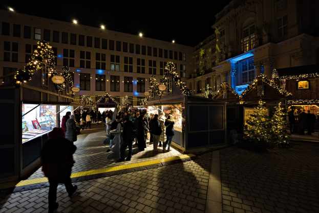 Ein geschäftiges Weihnachtsmarkt-Szenario bei Nacht mit Menschen um beleuchtete Stände, Stadtgebäude im Hintergrund und einem dunklen Himmel.