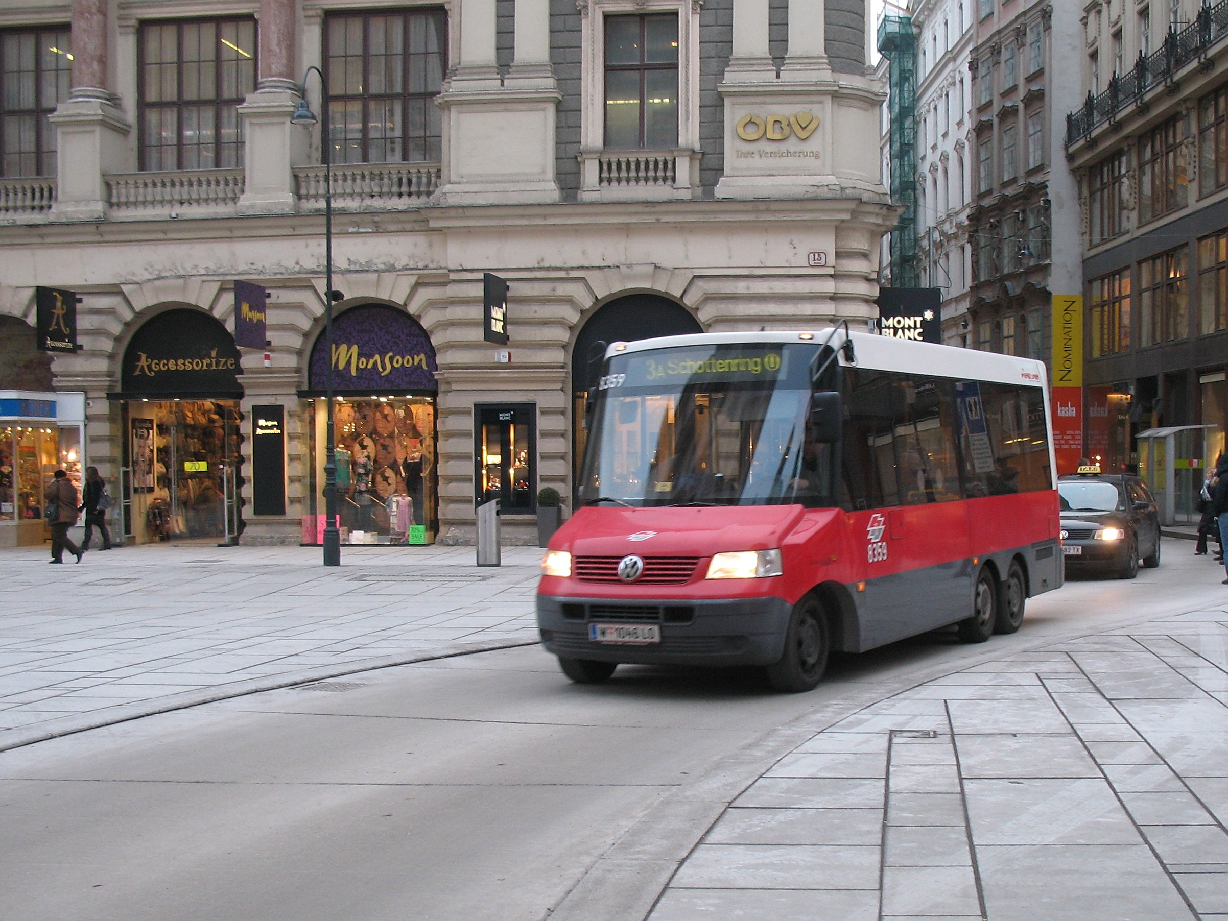Red shuttle bus driving down a city street with tall buildings, pedestrians on the sidewalk, light poles, and storefronts under a visible sky.