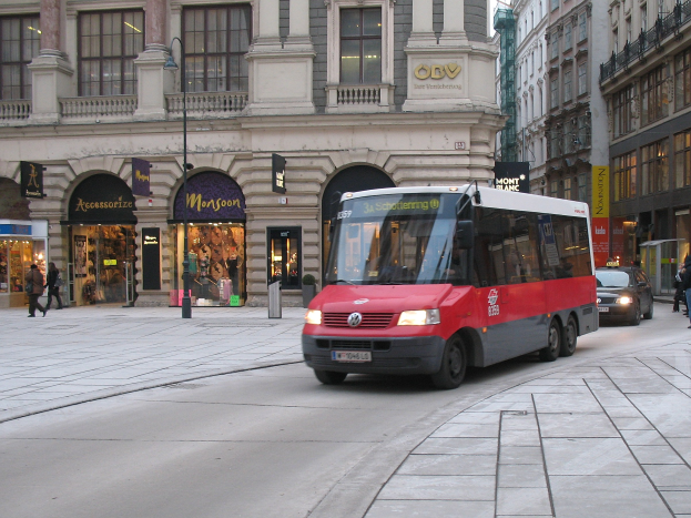 Red shuttle bus driving down a city street with tall buildings, pedestrians on the sidewalk, light poles, and storefronts under a visible sky.