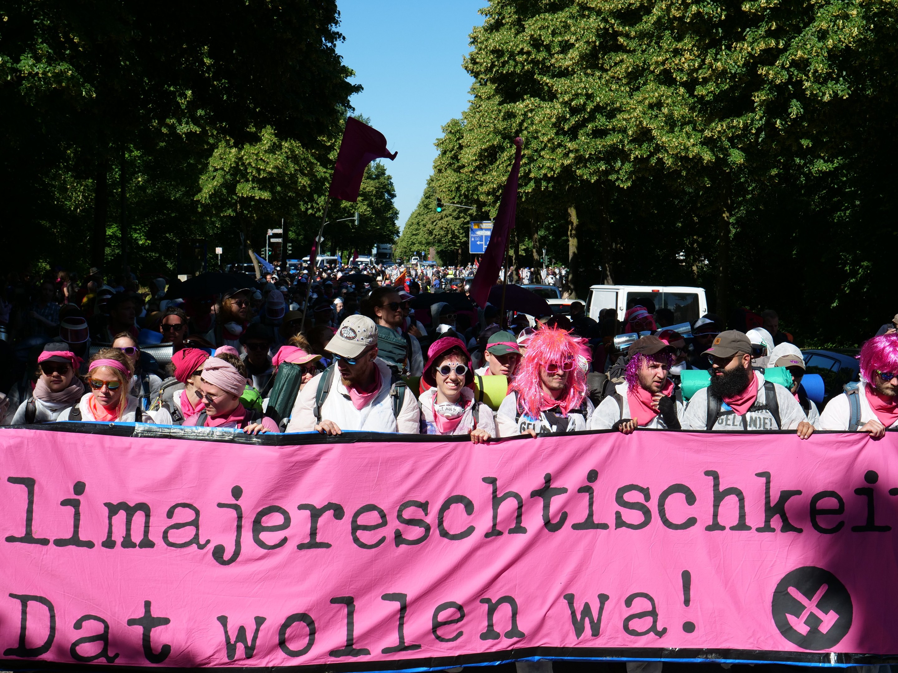 Gruppe von Menschen marschiert die Straße entlang und hält eine pinkfarbene Banner mit der Aufschrift 'Frauenmarsch in Berlin', wobei einige Mötze und Schutzbrillen tragen, andere Fahnen halten, vor dem Hintergrund von Bäumen, Fahrzeugen und Himmel.