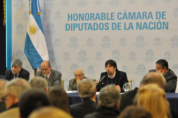 Eine Gruppe von Menschen sitzt in Reihen von St├╝hlen, mit einem Tisch vor der ersten Reihe, der einen Krug, Papiere und einen Stift enth├Ąlt. Eine Person in der Mitte spricht in ein Mikrofon, mit einer wei├čen und blauen Flagge dahinter und einem wei├čen und blauen Hintergrund, auf dem 'ehrenwerte kamara de' geschrieben steht.