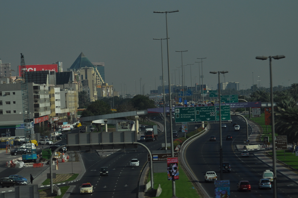 Eine Luftaufnahme einer Stadt mit Gebäuden, Türmen, Straßen, Kraftfahrzeugen, Werbetafeln, Schildern, Straßenlaternen, Bäumen, Menschen und Absperrgittern.