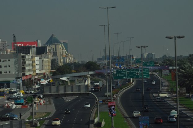 Eine Luftaufnahme einer Stadt mit Gebäuden, Türmen, Straßen, Kraftfahrzeugen, Werbetafeln, Schildern, Straßenlaternen, Bäumen, Menschen und Absperrgittern.