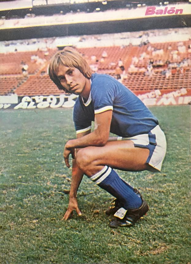 Ein Mann kniet auf einem Fußballfeld, umgeben von sitzenden Menschen und Stühlen mit Text darauf, trägt Schuhe.