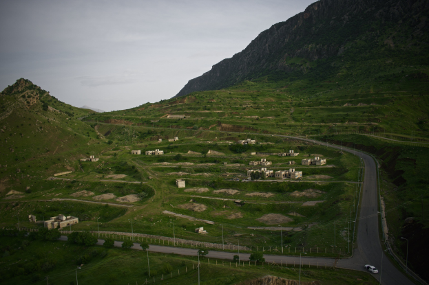 Ein kleines Dorf in einem Gebirgszug mit grünem Gras, Bäumen, Pfählen, Fahrzeugen auf einer Straße und einem sichtbaren Himmel.